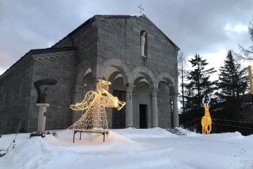 Chiesa di pietra in un paesaggio innevato e di fronte un angioletto e una renna fatti con le lucine natalizie