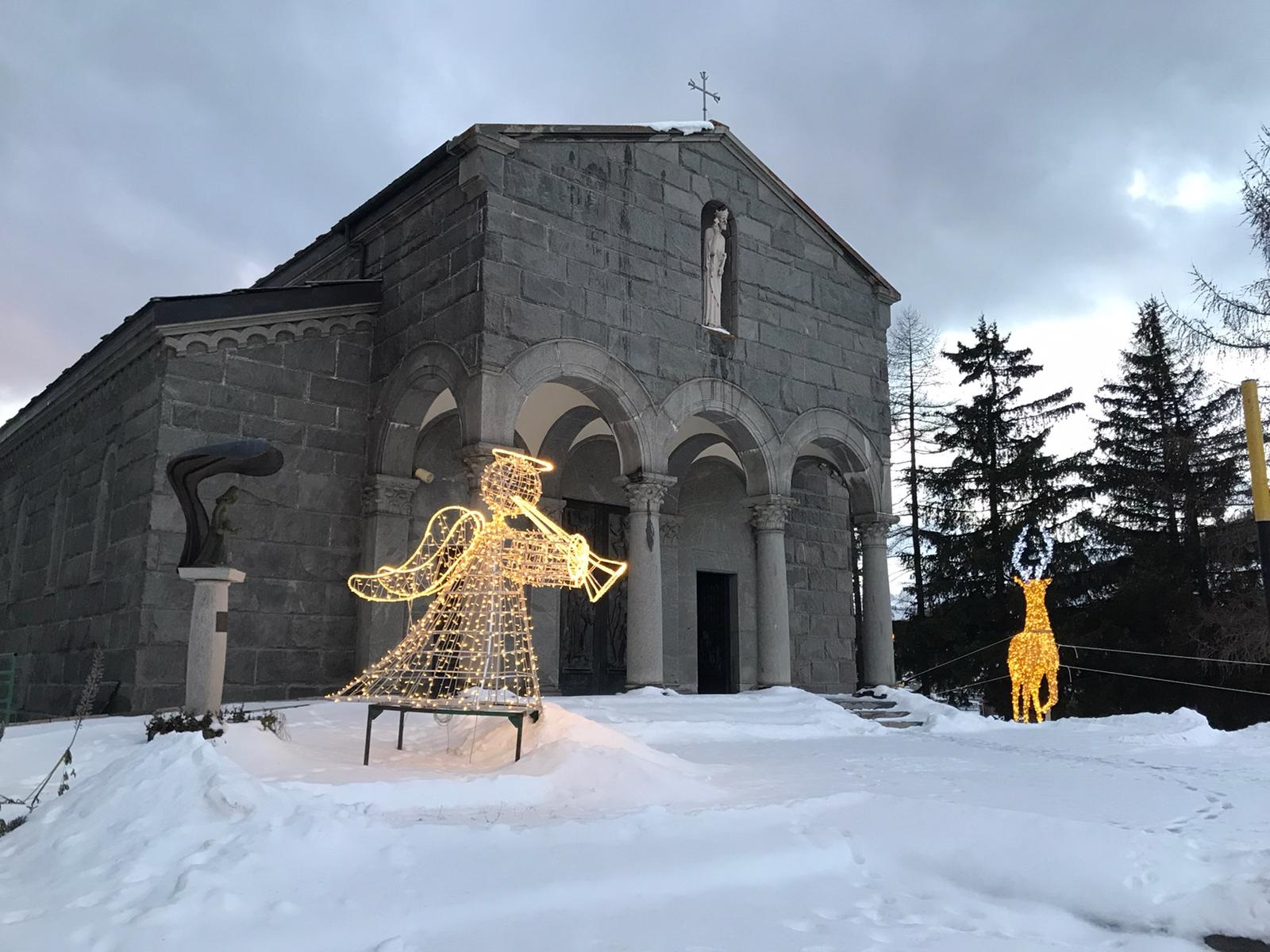 Chiesa di pietra in un paesaggio innevato e di fronte un angioletto e una renna fatti con le lucine natalizie